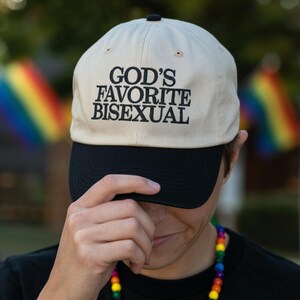 May include: A beige baseball cap with a black brim. The cap features the text "GOD'S FAVORITE BISEXUAL" in black block letters. The image is taken outdoors, with a person wearing the cap and a rainbow flag visible in the background.
