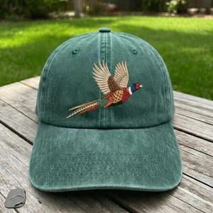 Puede incluir: Una gorra de b&eacute;isbol verde bosque con un dise&ntilde;o bordado de un fais&aacute;n en vuelo. El ave muestra un plumaje detallado en tonos marr&oacute;n, beige y azul iridiscente en la cabeza, centrado en el panel frontal de la gorra de seis paneles.