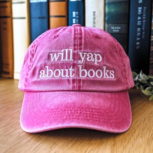 May include: A pink baseball cap with the embroidered text "will yap about books" in white. The cap is made of a soft fabric and has a curved brim. The background includes a collection of books.