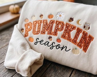 May include: A cream-colored sweatshirt featuring the embroidered text PUMPKIN season in orange glittery block letters and black cursive script. The design includes small embroidered icons of pumpkins, acorns, a white flower, a crescent moon, and an owl.