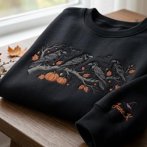 May include: A black crewneck sweatshirt featuring an embroidered design of four crows perched on a branch with orange pumpkins, autumn leaves, and small bats. The sleeve is embroidered with the name Jane.K and a small purple witch hat icon.