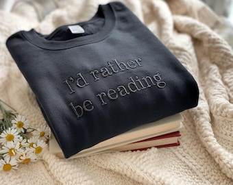 May include: A dark gray sweatshirt with the embroidered text "I'd rather be reading." The sweatshirt is folded on top of a stack of books with red covers. The background includes a cream-colored knitted blanket and a few daisies.