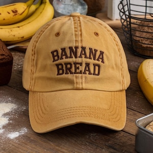 May include: Mustard-colored baseball cap with the words "BANANA BREAD" embroidered in brown. The hat has a curved brim and a classic six-panel design. The hat is made of a soft, worn material.