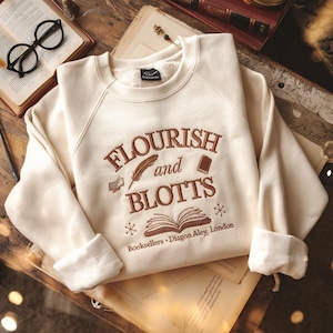 May include: Cream-colored sweatshirt with the embroidered text "FLOURISH and BLOTTS Booksellers Diagon Alley, London." The design includes a feather, books, and snowflakes in brown. The sweatshirt has a crew neck and cuffed sleeves.