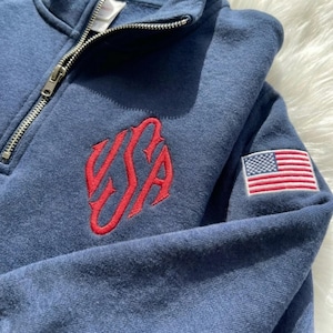 May include: Navy blue sweatshirt with a quarter-zip neckline. Features a red embroidered "USA" design on the front and a small American flag patch on the sleeve. The sweatshirt is made of a soft, comfortable material.