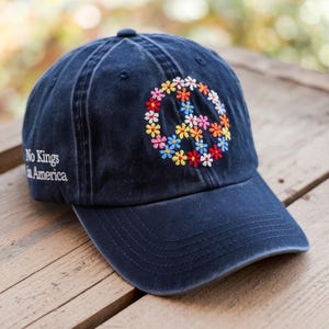 May include: A navy blue baseball cap with a floral peace sign embroidered on the front in various colors. The text "No Kings in America" is embroidered on the side. The cap is made of a soft, worn material.