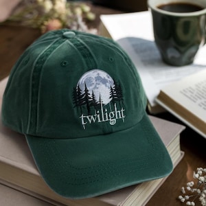 May include: A dark green baseball cap with a curved brim. The cap features an embroidered design of a full moon, pine trees, and the word "twilight" in white script. The cap is sitting on top of a stack of books.