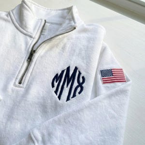 May include: White quarter-zip sweatshirt with a navy blue monogram and an American flag patch on the sleeve. The sweatshirt has a silver zipper and a soft, textured appearance. The monogram features the letters "MM8".
