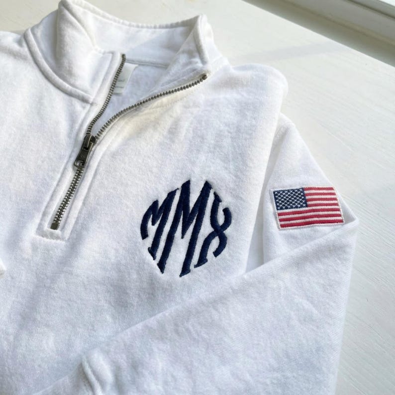 May include: White quarter-zip sweatshirt with a navy blue monogram and an American flag patch on the sleeve. The sweatshirt has a silver zipper and a soft, textured appearance. The monogram features the letters "MM8".