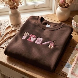 May include: A dark brown crewneck sweatshirt featuring embroidered pink kitchen-themed icons across the chest, including a utensil holder, a stand mixer, a bag labeled flour, a rolling pin, and a set of measuring cups arranged in a horizontal line.