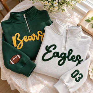 May include: A dark green quarter-zip sweatshirt with yellow Bears text and a football patch on the sleeve, alongside a light gray quarter-zip sweatshirt with dark green Eagles text and the number 03 on the sleeve.