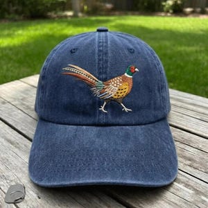 Puede incluir: Una gorra de b&eacute;isbol azul marino con un dise&ntilde;o bordado de un fais&aacute;n en el panel frontal. El ave aparece de perfil con la cabeza verde, un anillo blanco en el cuello y plumas con patrones en tonos marr&oacute;n, beige y negro en el cuerpo.