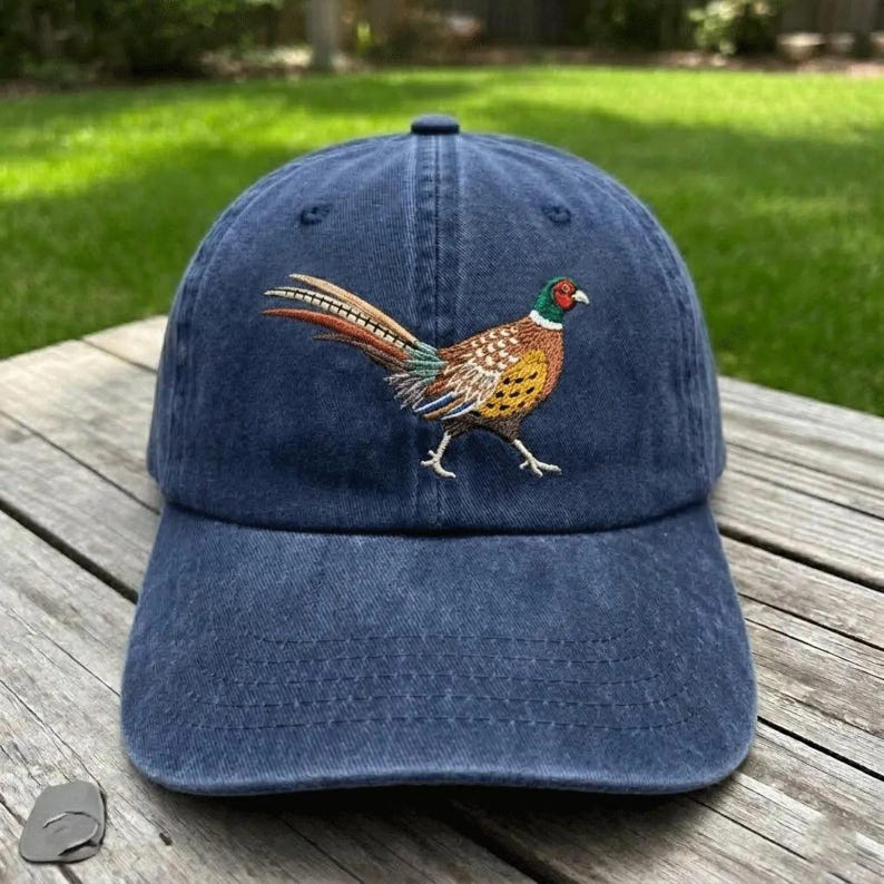 Puede incluir: Una gorra de b&eacute;isbol azul marino con un dise&ntilde;o bordado de un fais&aacute;n en el panel frontal. El ave aparece de perfil con la cabeza verde, un anillo blanco en el cuello y plumas con patrones en tonos marr&oacute;n, beige y negro en el cuerpo.