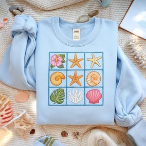 May include: Light blue sweatshirt featuring a grid of embroidered sea-themed designs. The grid includes a pink hibiscus flower, starfish, seashells, and coral. The sweatshirt is made of a soft material and has a crew neck.