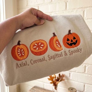 May include: A beige sweatshirt featuring an embroidered design of four orange pumpkins in various cross-sections and a jack-o'-lantern face. Below the pumpkins, the text "Axial, Coronal, Sagittal & Spooky" is embroidered in a dark brown thread.