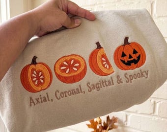 Może przedstawiać: Beżowa bluza z wyhaftowanym wzorem czterech pomarańczowych dyń w różnych przekrojach oraz twarzą jack-o'-lantern. Pod dyniami wyhaftowano ciemnobrązową nicią napis "Axial, Coronal, Sagittal & Spooky".