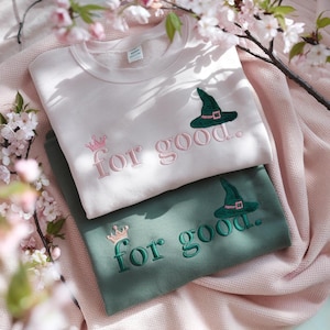 May include: Two sweatshirts, one light pink and one green, are stacked on a pink blanket. The light pink sweatshirt has the phrase "for good." embroidered in pink, a pink crown, and a green witch hat. The green sweatshirt has the same design in green.