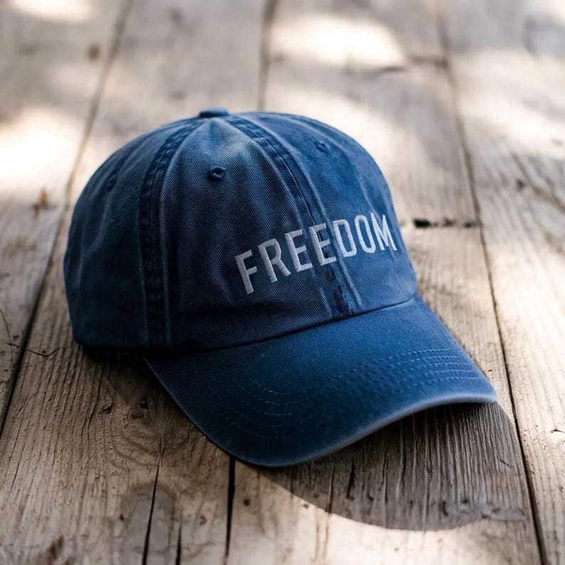 Puede incluir: Gorra de b&eacute;isbol azul marino con la palabra "FREEDOM" bordada en blanco. La gorra est&aacute; hecha de un material suave y tiene una visera curva. El sombrero est&aacute; sobre una superficie de madera.