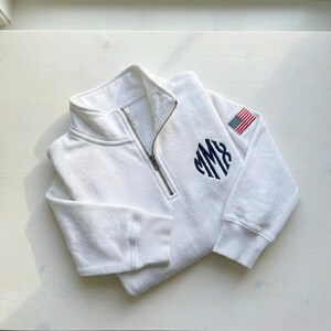 May include: White zip-up sweatshirt with a navy blue monogram and an American flag patch on the sleeve. The sweatshirt has a high collar and a silver zipper. The monogram is in a circular design.