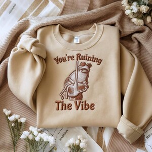 May include: Beige crewneck sweatshirt with the embroidered phrase "You're Ruining The Vibe" and a frog illustration. The frog is brown and appears to be holding a pole. The sweatshirt is folded on a beige blanket.