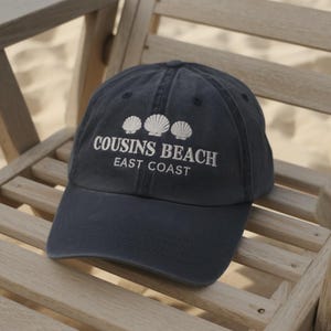 May include: Navy blue baseball cap with the text "COUSINS BEACH EAST COAST" embroidered in white. The cap features three white seashell designs above the text. The hat is resting on a wooden chair.