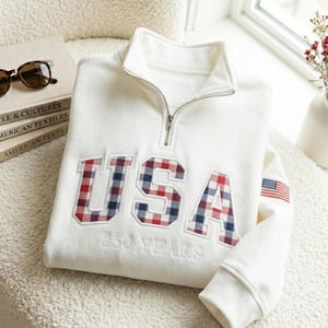 May include: A white sweatshirt with a quarter-zip collar and the word "USA" in red, white, and blue plaid letters. The number "250 YEARS" is embroidered below the letters. An American flag patch is on the sleeve.