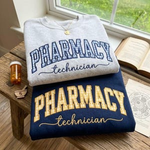 May include: A light gray crewneck sweatshirt with blue gingham PHARMACY technician text and a dark navy sweatshirt with yellow gingham PHARMACY technician text. Both feature embroidered lettering in a collegiate font style.