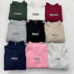 May include: Nine different colored quarter-zip sweatshirts are displayed. Colors include forest green, white, maroon, oxford gray, black, sand, navy, pink, and ash gray. Each sweatshirt has a zipper and the color name printed on it.