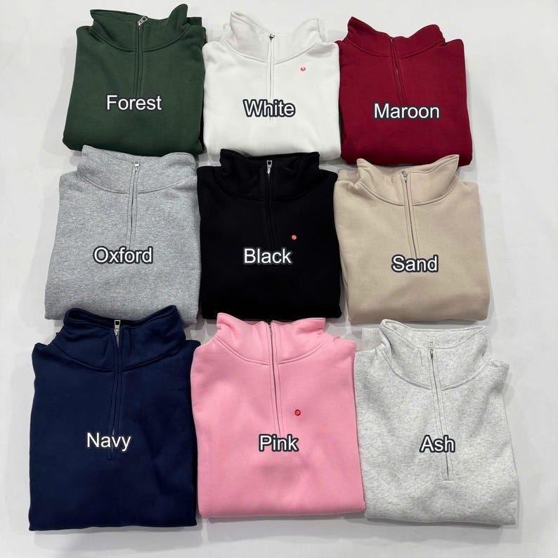 May include: Nine different colored quarter-zip sweatshirts are displayed. Colors include forest green, white, maroon, oxford gray, black, sand, navy, pink, and ash gray. Each sweatshirt has a zipper and the color name printed on it.