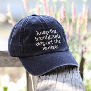 May include: A navy blue baseball cap with the embroidered text "Keep the immigrants deport the racists" in white. The cap is made of a soft fabric and has a curved brim. The hat is sitting on a wooden surface.