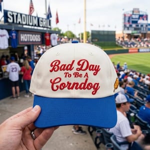 May include: A baseball cap with a white crown and a blue brim. The cap features the embroidered phrase "Bad Day To Be A Corndog" in red script, along with a small corndog graphic. The cap is being held in front of a stadium.