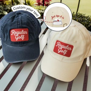 May include: Two baseball caps, one navy blue and one cream-colored. Both caps feature a red rectangular patch with the words "Regular Golf" in white. The navy cap has the text "Custom your text" above it, and the cream cap has "just tap it in" on the back.