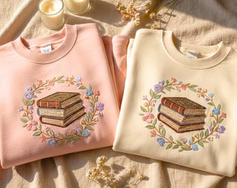 May include: Two sweatshirts, one light pink and one cream, are displayed. Each features an embroidered design of a stack of books encircled by a floral wreath. The sweatshirts are on a beige surface, with two lit candles and dried flowers.