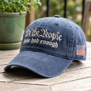 May include: A navy blue baseball cap with the embroidered text "We the People have had enough" in white script. The cap features a small American flag patch on the side. The hat is made of a soft, worn fabric.