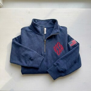 May include: A navy blue quarter-zip sweatshirt with a stand-up collar. The sweatshirt features a red embroidered "USA" design on the chest and a small American flag patch on the sleeve. The zipper is silver.