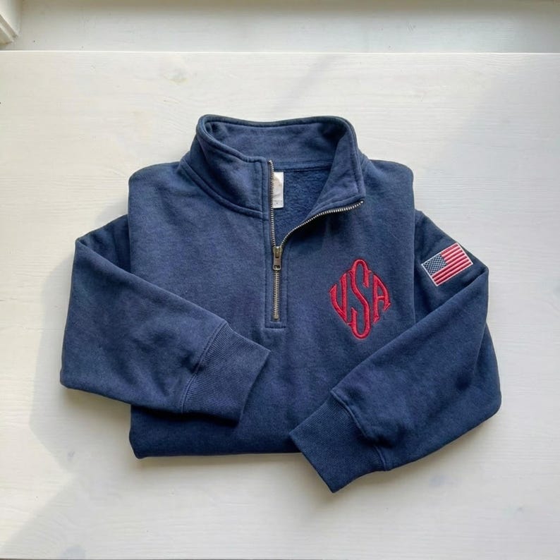 May include: A navy blue quarter-zip sweatshirt with a stand-up collar. The sweatshirt features a red embroidered "USA" design on the chest and a small American flag patch on the sleeve. The zipper is silver.