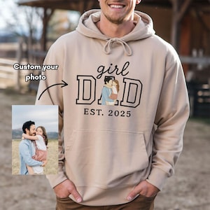 May include: A beige hoodie with a front pocket and drawstring. The front features the text "girl DAD" in a bold, black font, with an illustration of a father and daughter. Below is "EST. 2025". A photo can be customized.