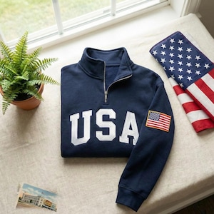 May include: Navy blue sweatshirt with a quarter-zip collar and the word "USA" in large white letters. An American flag patch is on the left sleeve. An American flag is folded nearby.