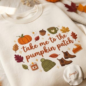 May include: A cream-colored sweatshirt featuring embroidered autumn-themed icons including a pumpkin, acorn, mug, leaves, pie slice, boots, candle, and beanie. The text "take me to the pumpkin patch" is centered. A sleeve cuff shows the name "Janey.S".