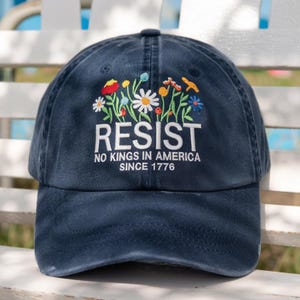 May include: A navy blue baseball cap with embroidered floral design and the word "RESIST" in white. The text "NO KINGS IN AMERICA SINCE 1776" is below the word "RESIST".