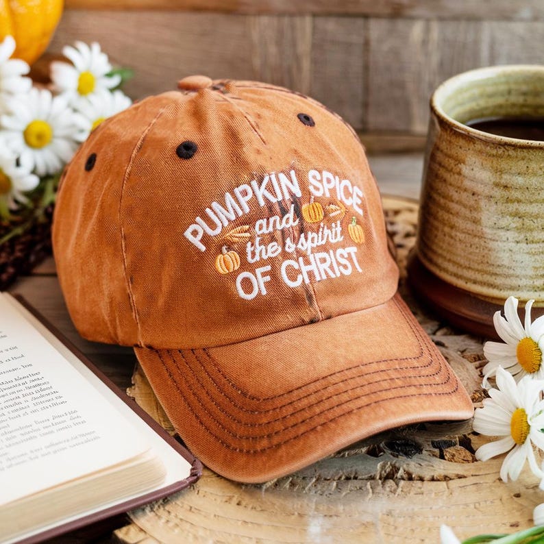 May include: A burnt orange baseball cap with the text "PUMPKIN SPICE and the spirit OF CHRIST" in white. The cap features a curved brim and black eyelets. The cap is sitting on a wooden surface.
