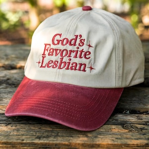 May include: A baseball cap with a cream-colored crown and a maroon brim. The phrase "God's Favorite Lesbian" is embroidered in red on the front of the cap. The cap has a maroon button on top.