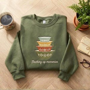 May include: An olive green sweatshirt featuring an embroidered design of stacked, vintage-style mixing bowls in shades of blue, yellow, orange, and green. The text "Stacking up memories" is below the bowls. A mug of coffee and a potted plant are in the background.