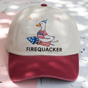 May include: Cream-colored baseball cap with a maroon brim and button. The cap features an embroidered design of a duck wearing a patriotic shirt and a party hat, riding a firework. The word "FIREQUACKER" is embroidered below the duck.
