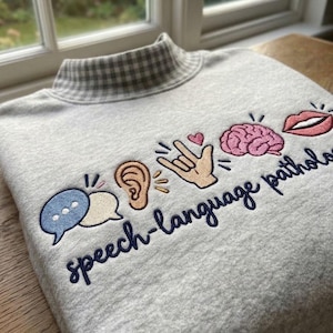 May include: A light gray sweatshirt with a mock neck featuring a gray and white gingham pattern. The front is embroidered with colorful icons including speech bubbles, an ear, a hand sign, a brain, and lips, with the text speech-language pathology.