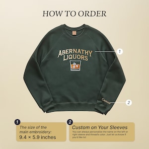Dark green embroidered sweatshirt featuring the words "Abernathy Liquors" in gold and cream thread, with a stitched image of a whiskey glass containing amber liquid and ice cubes. Inspired by Haymitch Abernathy from The Hunger Games, this design subtly references the character’s iconic drink.