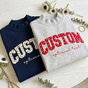 May include: A navy blue sweatshirt and a heather gray sweatshirt, both featuring a mock neck and the word CUSTOM in large, polka-dot applique letters, with the words optional text embroidered in cursive underneath the main lettering.