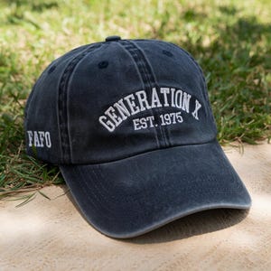 May include: A navy blue baseball cap with the text "GENERATION X EST. 1975" in white, arched across the front. The letters "FAFO" are embroidered on the side. The cap is sitting on a light-colored surface with green grass in the background.
