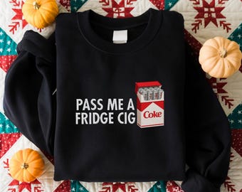 May include: Black sweatshirt with the text "PASS ME A FRIDGE CIG" in white. A graphic of a cigarette pack with the "Coke" logo is on the right. The sweatshirt is surrounded by small pumpkins on a patterned quilt.