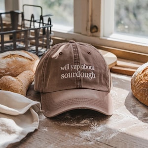 May include: A brown baseball cap with the embroidered text "will yap about sourdough" in white. The cap is on a wooden surface with loaves of bread and a white cloth. The cap has a curved brim and a classic six-panel design.